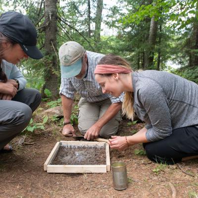 Group of researchers examining a soil sample in a forested area, using a wooden frame and metal can, while kneeling on the ground and focusing on their outdoor fieldwork.